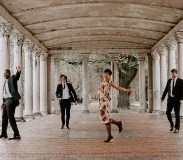 Four people in formal wear walking through a white-columned stone arcade, one mid-dance.