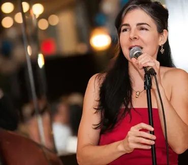 Singer in a red dress performing into a microphone at a live event, with blurred lights in the background