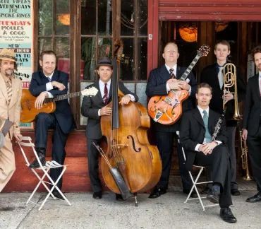 Six-piece jazz band posing outside a café, holding guitars, upright bass, trumpet, and trombone.