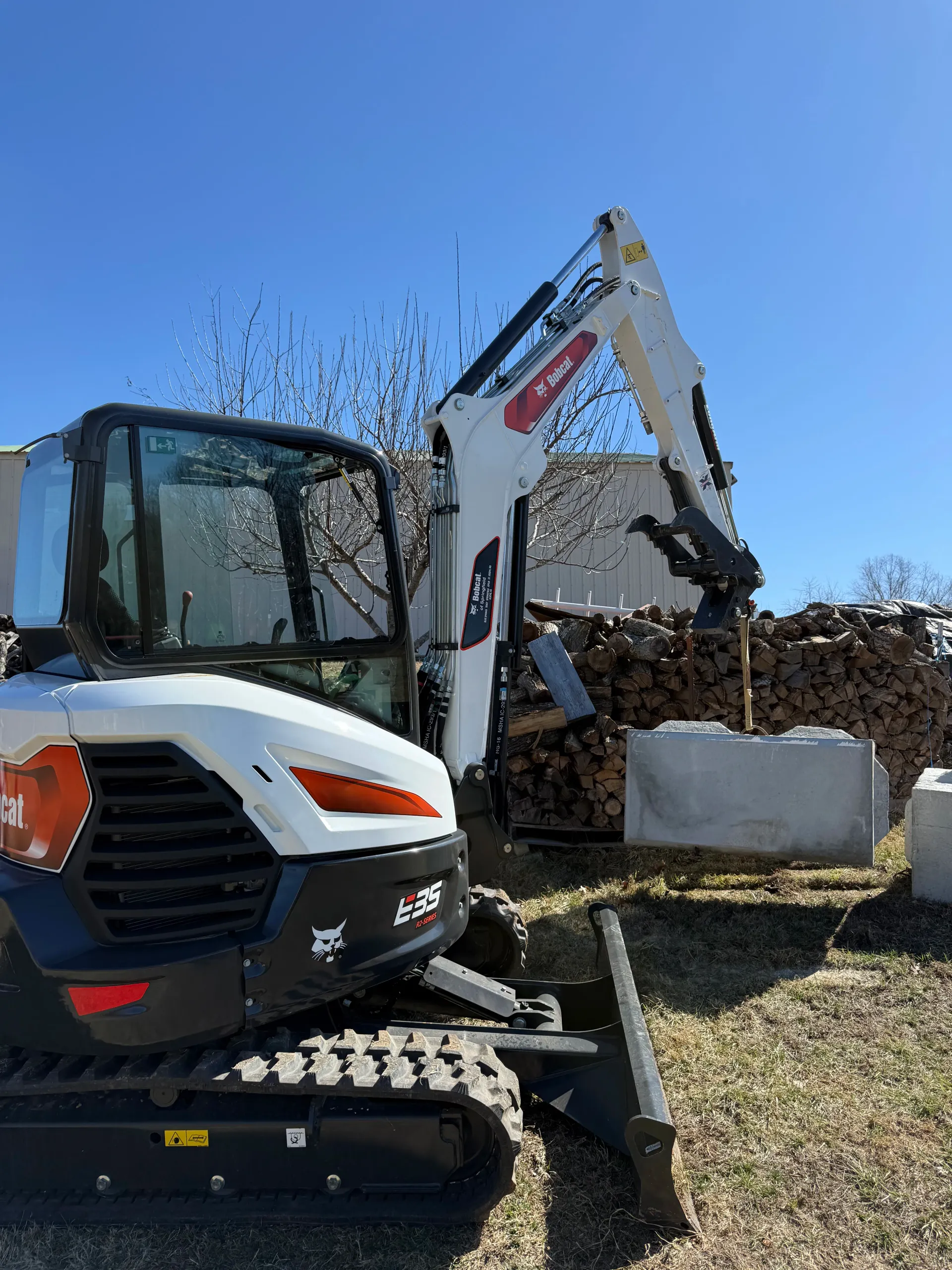 A white and black Bobcat E29 compact excavator lifts a concrete barrier in an outdoor setting on a sunny day.