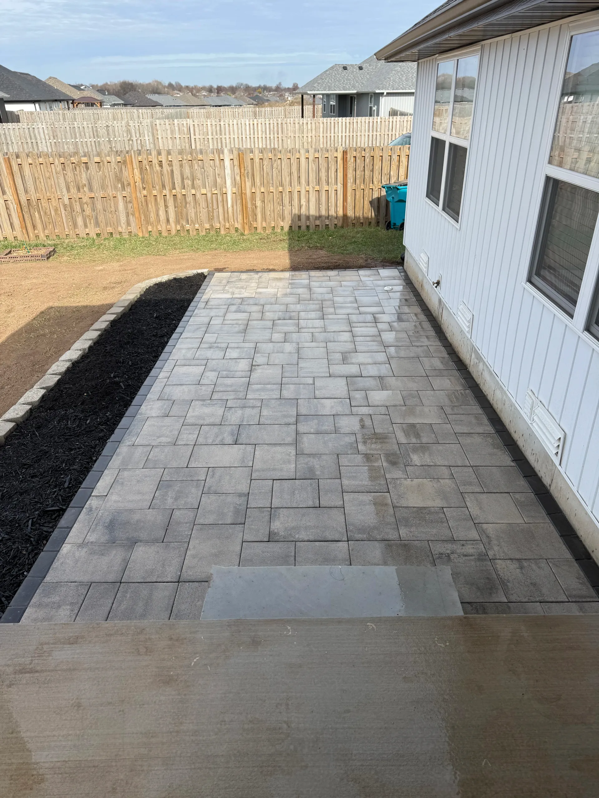 A backyard patio made of grey stone pavers with a black mulch border, adjacent to a white house wall.