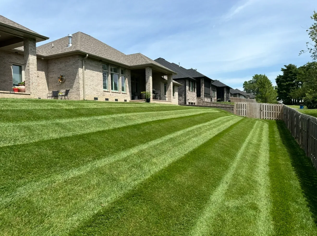 A row of beige brick houses with backyards featuring neatly mowed grass with alternating light and dark green stripes.
