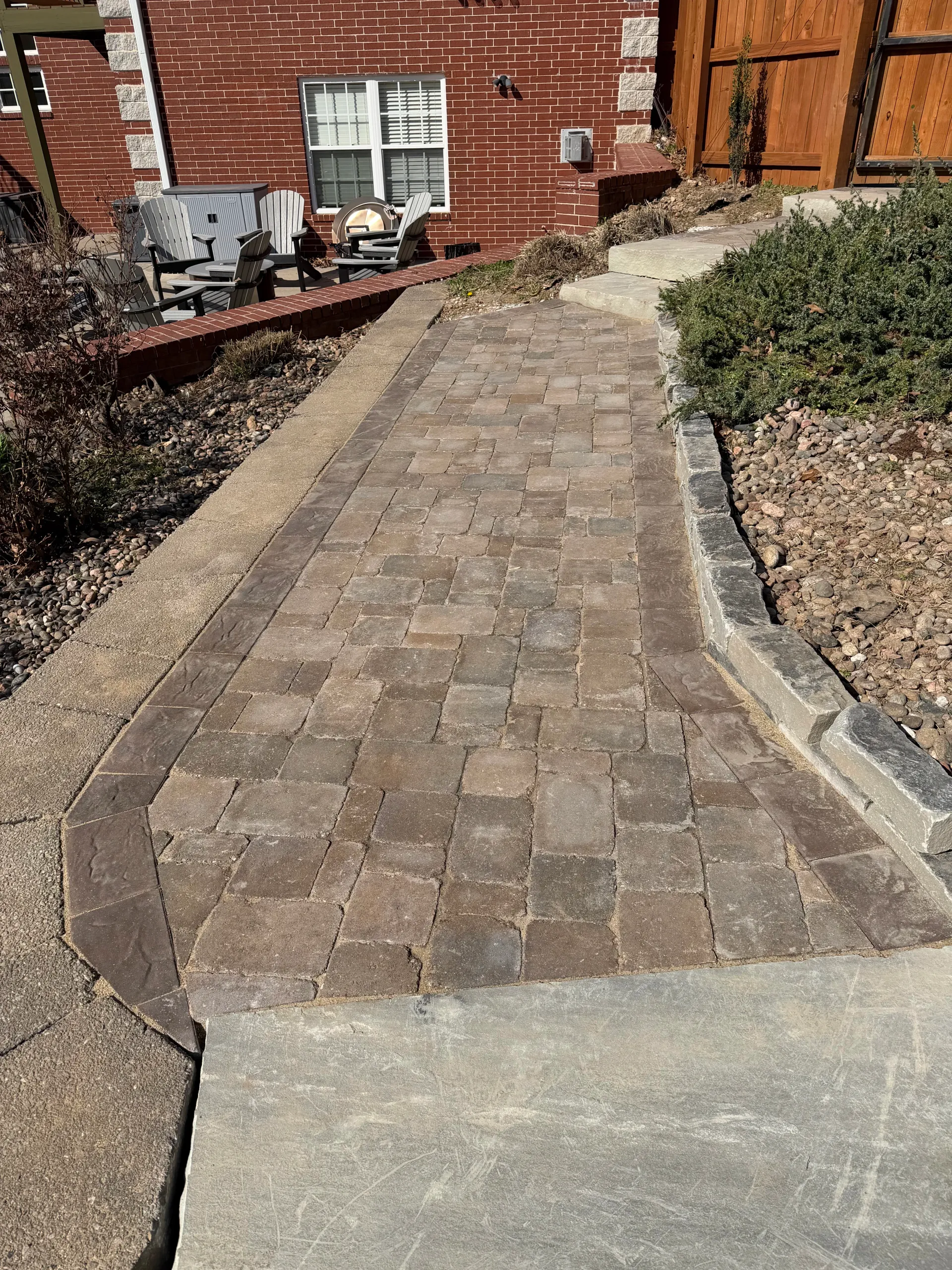 A brick walkway transitions from a concrete sidewalk toward a brick house with a wooden fence.