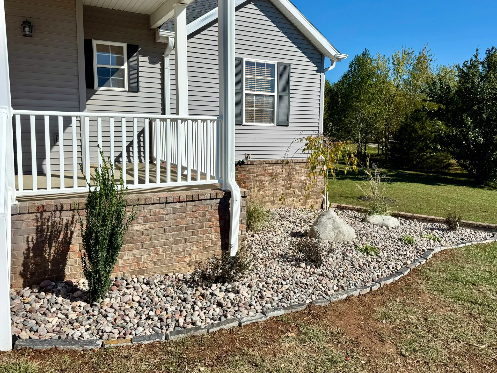 A light-grey house porch with white railings, a stone foundation, and a garden bed filled with river rocks.