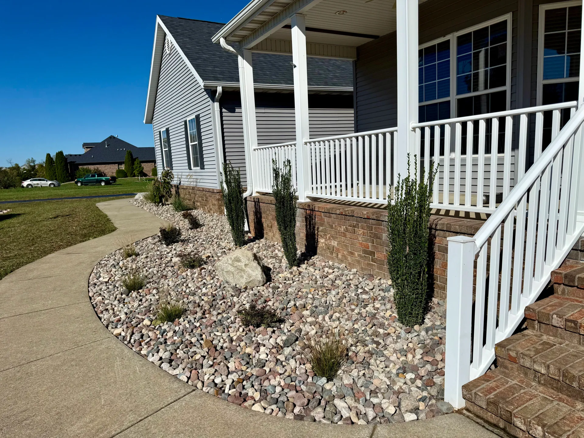 A view of a house porch with white railings, brick foundation, and a landscaped rock garden bed with small green shrubs.