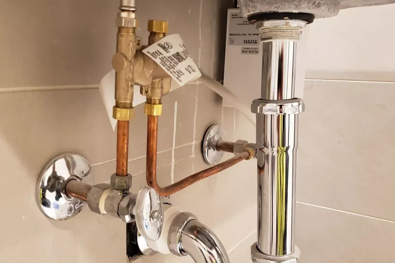 Plumbing under a sink with copper pipes, chrome valves, and a shiny drain pipe against tiled walls