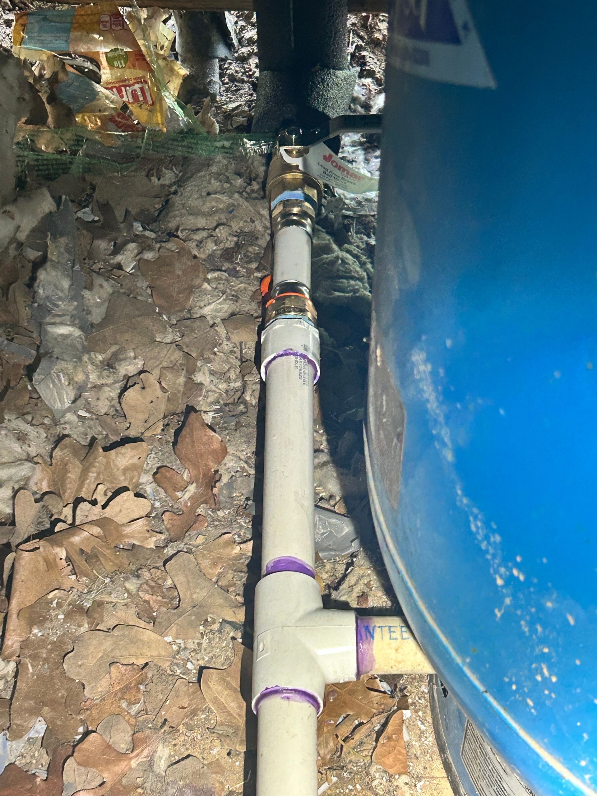 PVC pipes connected to a blue water tank with a T-joint and metal valve, set against a rough, brick-like wall.