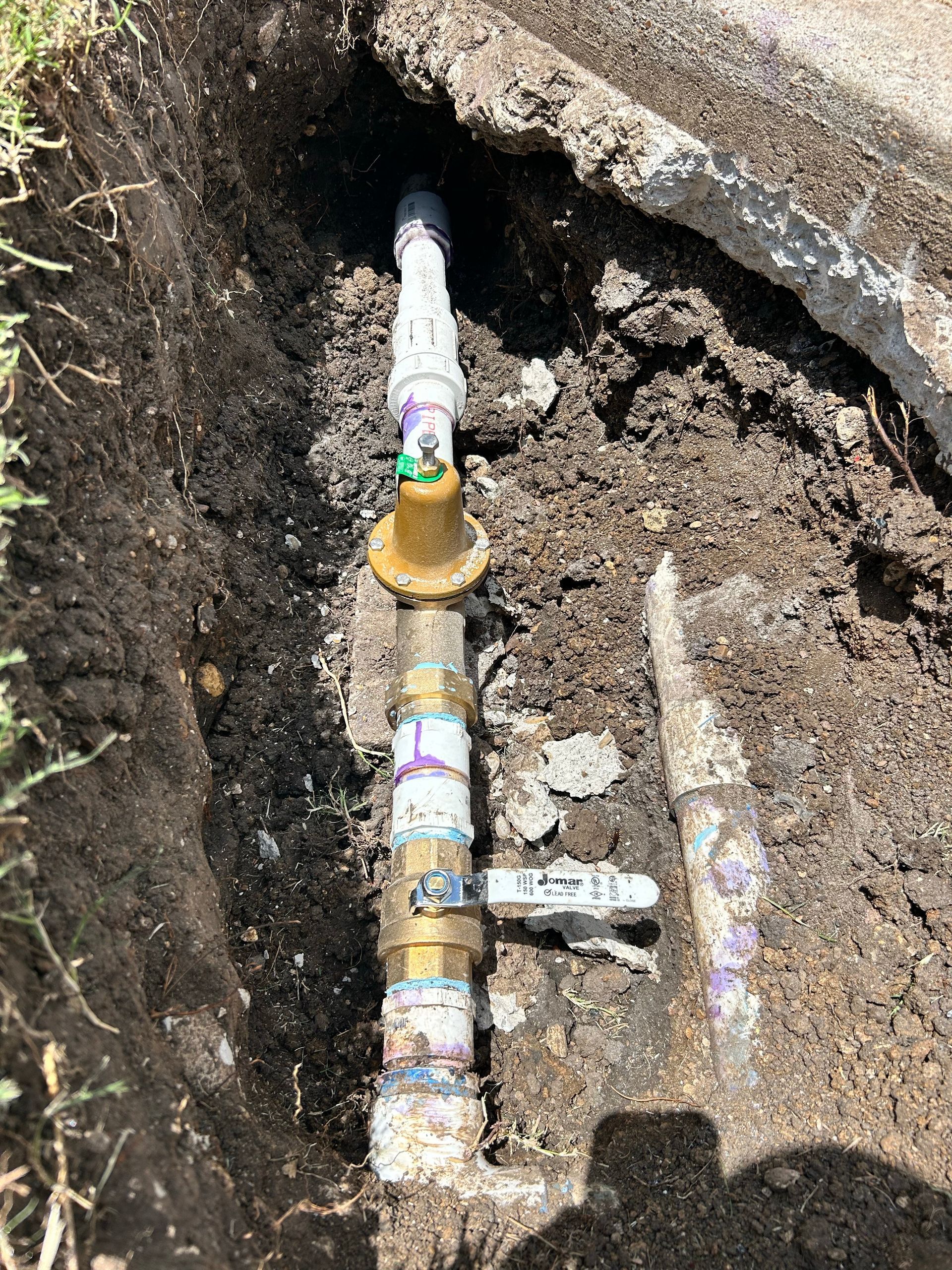 A trench with buried PVC and brass water pipes and a shut-off valve, dug in the dirt.