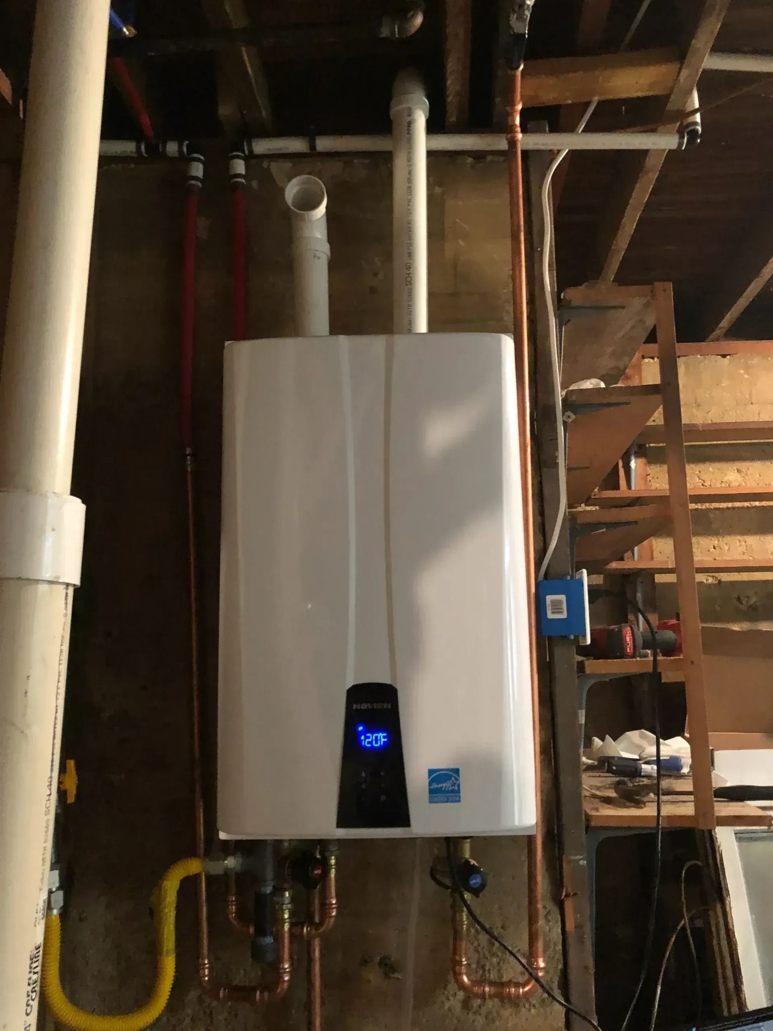 White tankless water heater on a concrete wall in a basement, with associated pipes and vents.