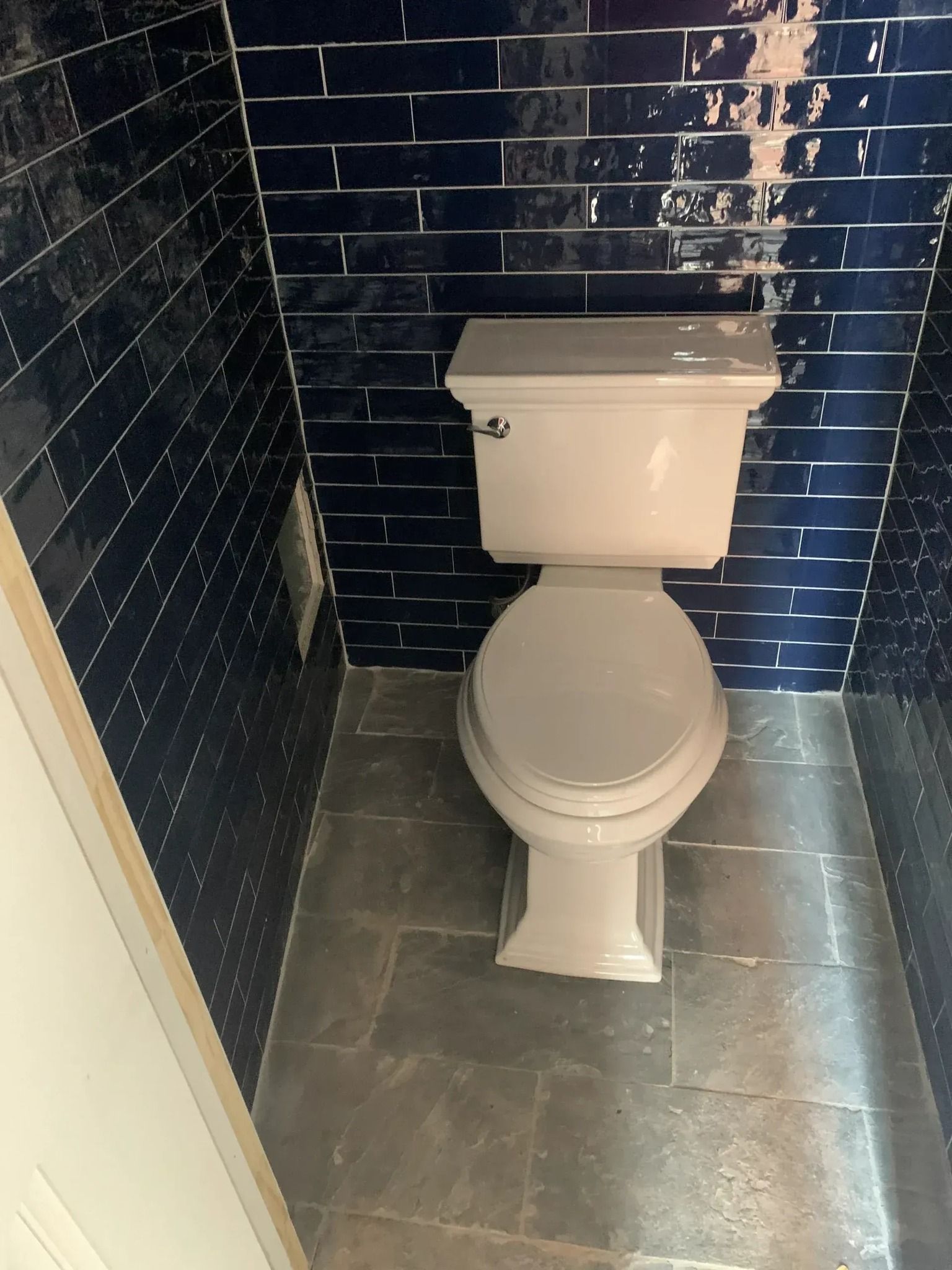 A toilet in a small bathroom with dark blue tiled walls and gray tiled floor.
