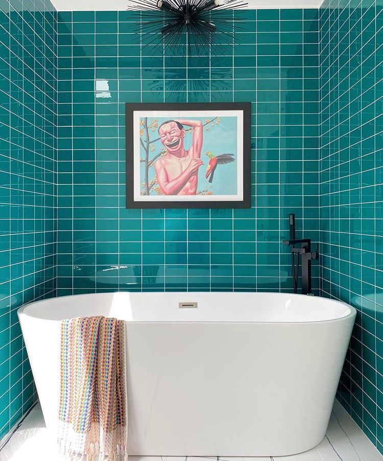 White bathtub against teal tiled wall with art and black faucet. Towel draped on tub.