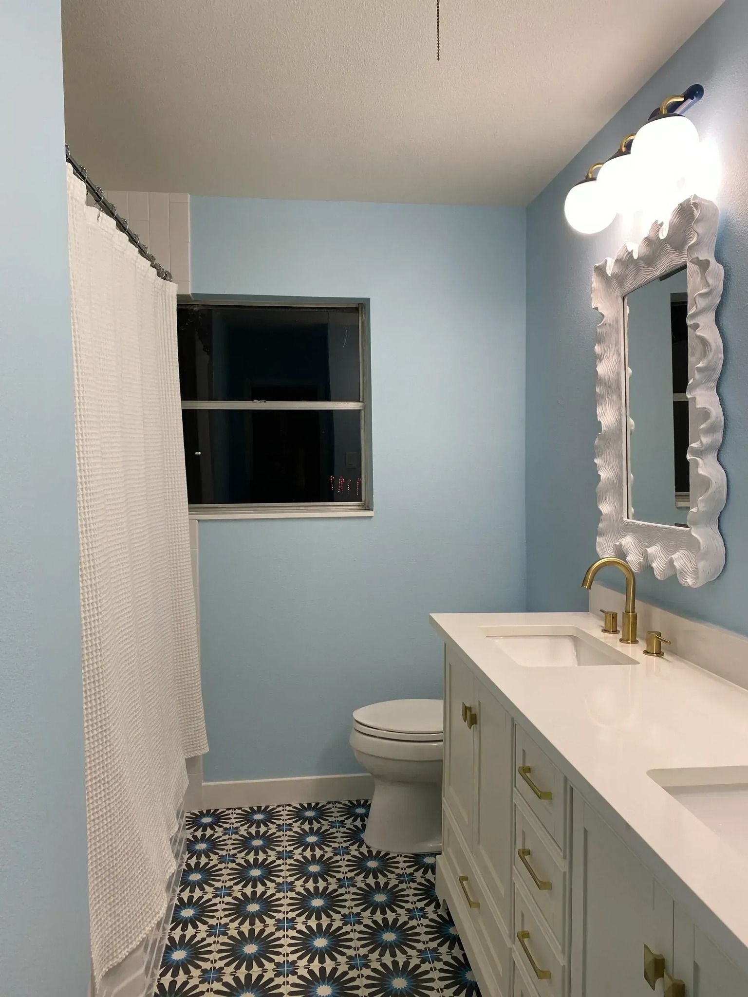 Blue bathroom with patterned floor, white vanity with gold fixtures, ornate mirror, and window.