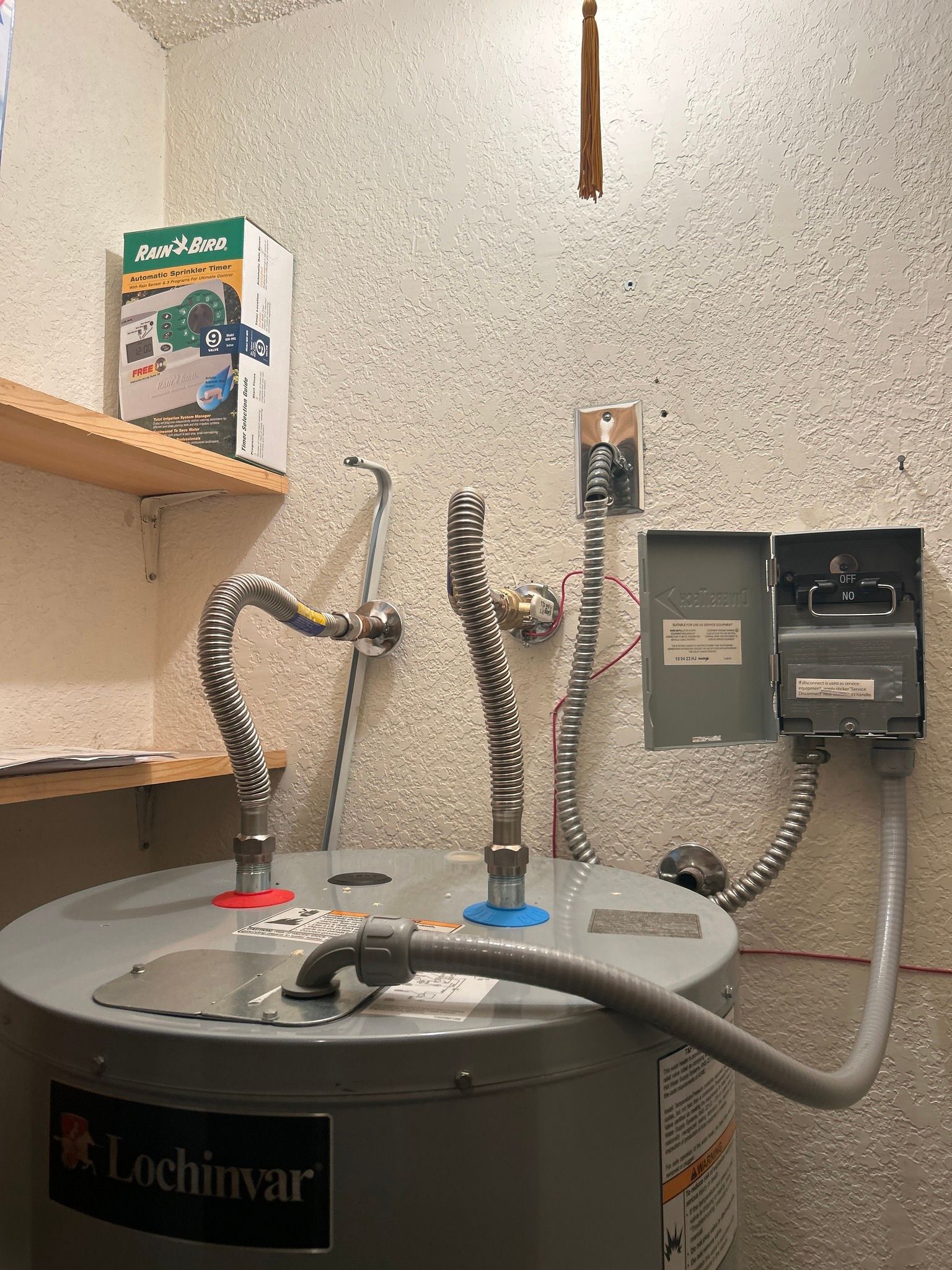 Water heater with flexible metal pipes and electrical components in a speckled wall setting.