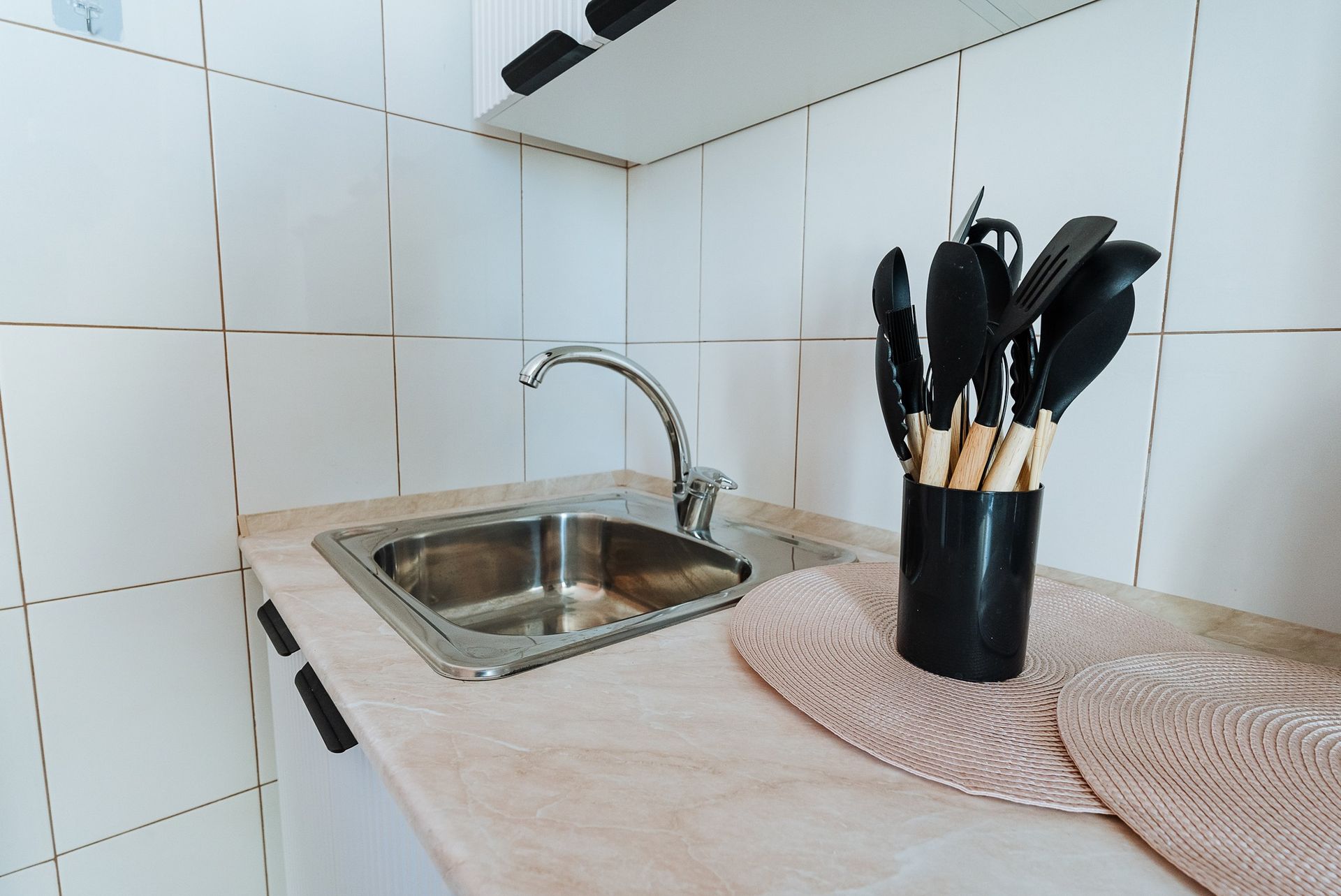 Kitchen sink with utensils in a holder on a countertop. Beige and white tiles surround.