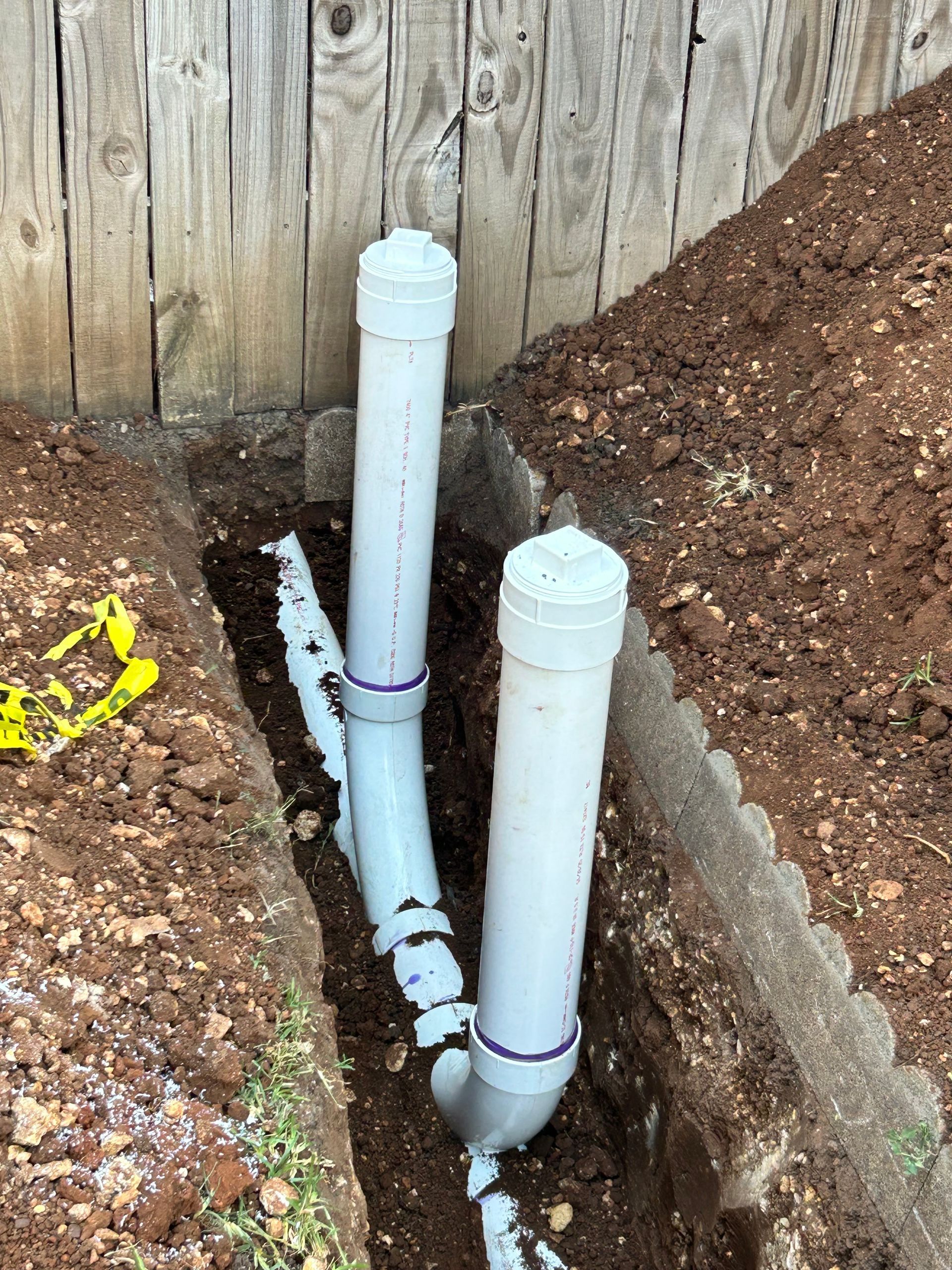 PVC pipes in a trench, next to a wooden fence. Brown soil surrounds the pipes.