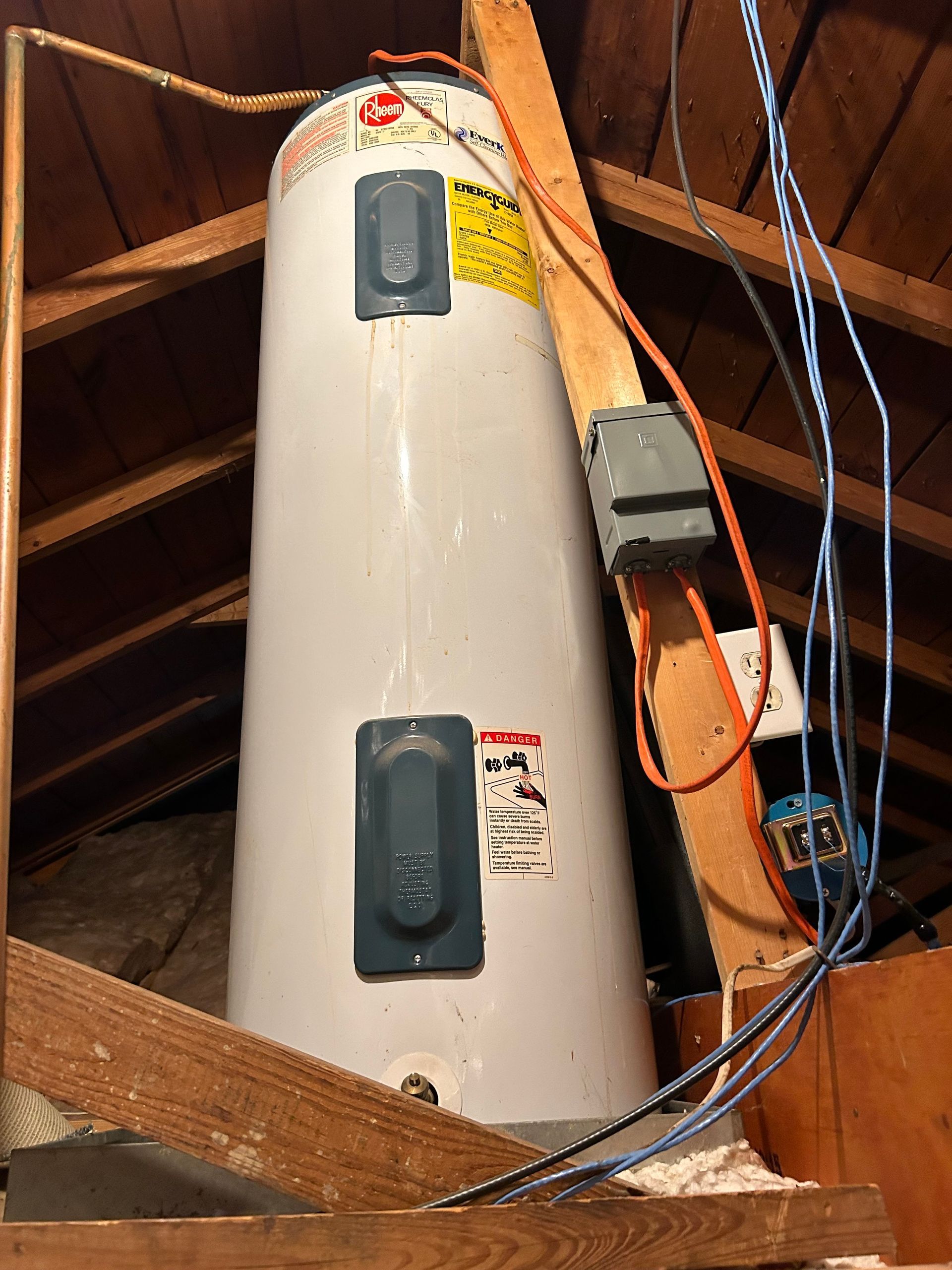 A white water heater in a wooden attic, with electrical wiring and a support structure.