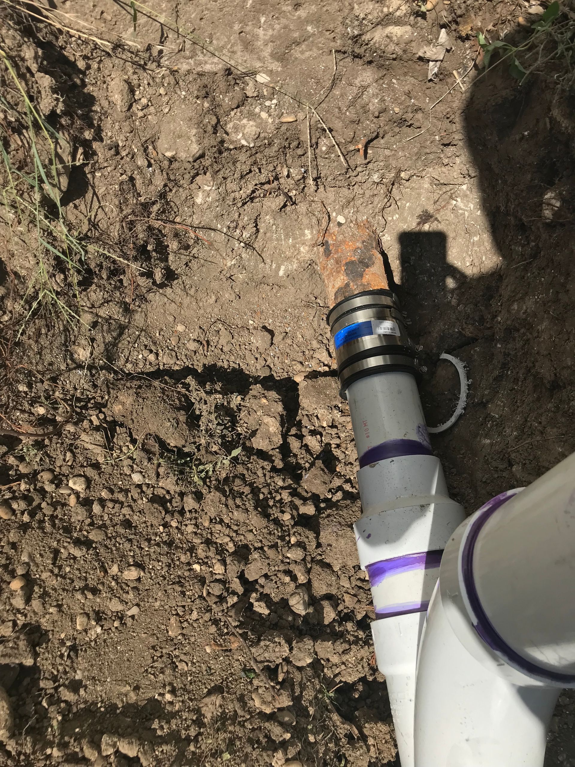 White PVC pipes connected to a brown metal pipe in a dirt setting. Black and blue connectors are visible.