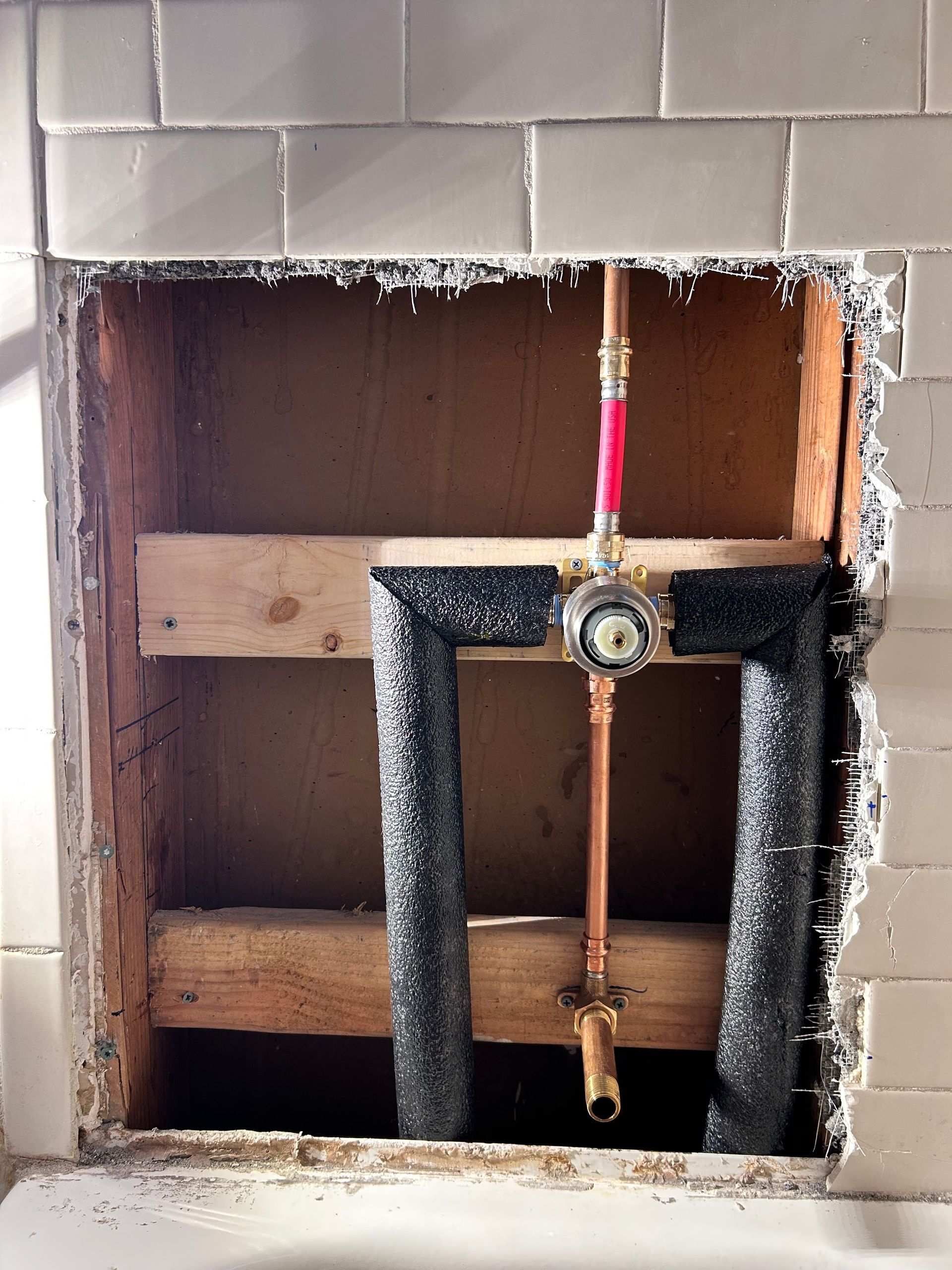 Hole in a tiled wall exposing copper plumbing and wooden supports. Insulation surrounds the pipes.