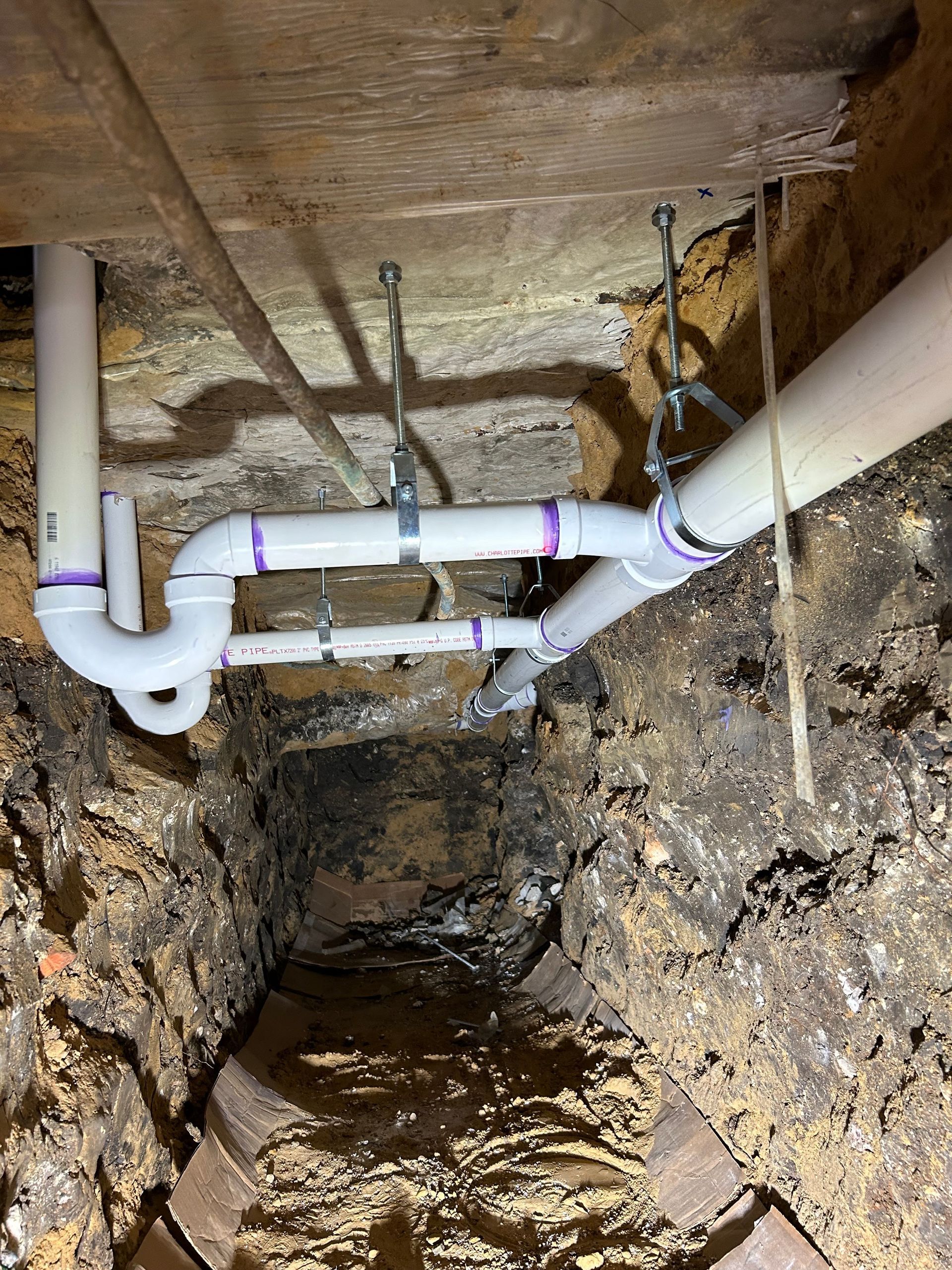 White PVC plumbing pipes installed in a trench, supported by metal hangers.