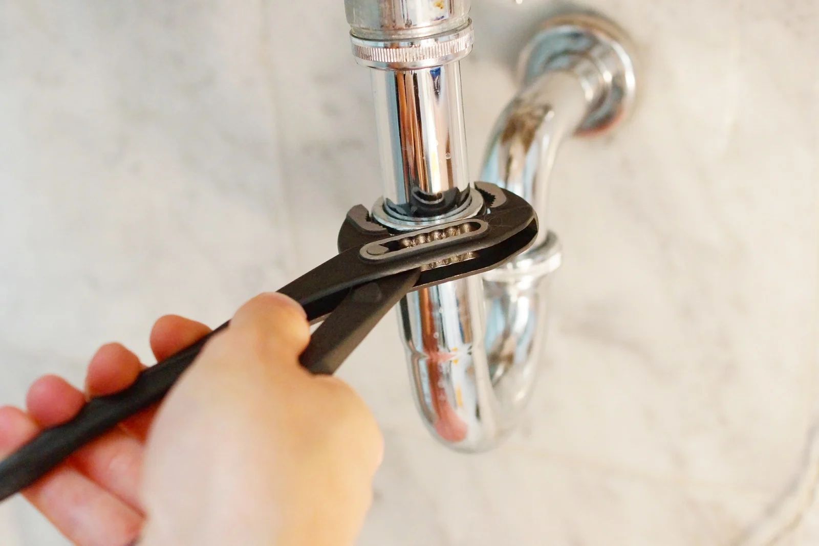 Hand using a wrench to tighten a chrome pipe under a sink.