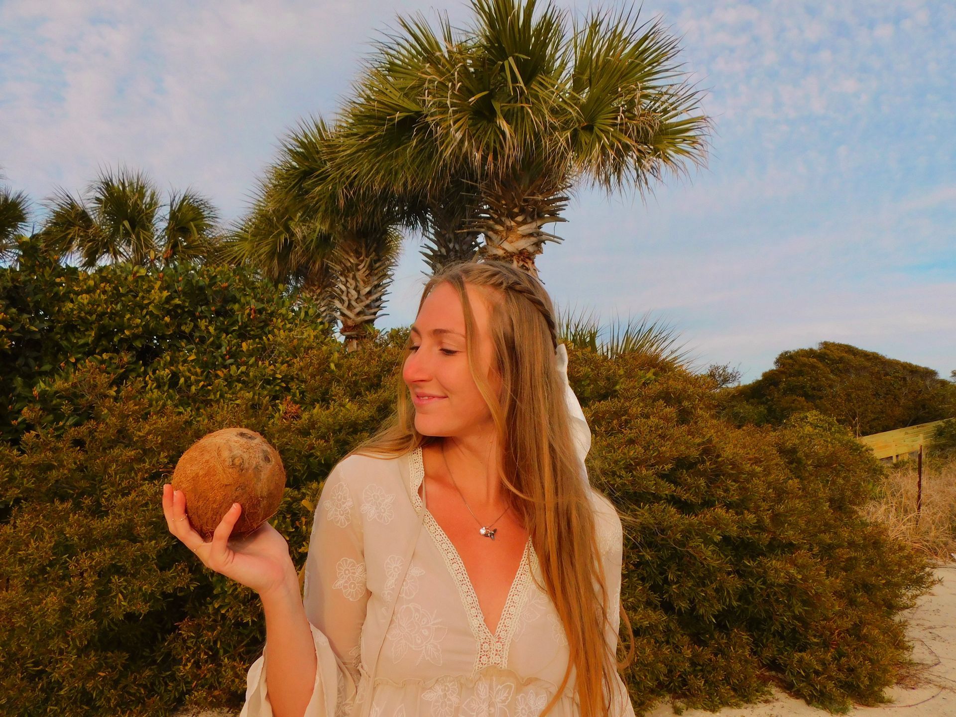 Woman with long blonde hair, holding a coconut, smiles outdoors near palm trees and green bushes.