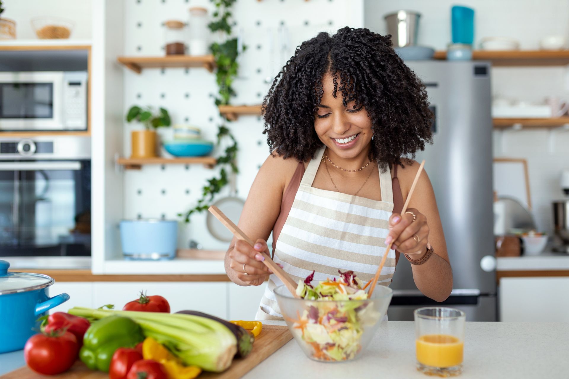 Woman making a salad in a light, modern kitchen, smiling as she mixes ingredients with wooden spoons.