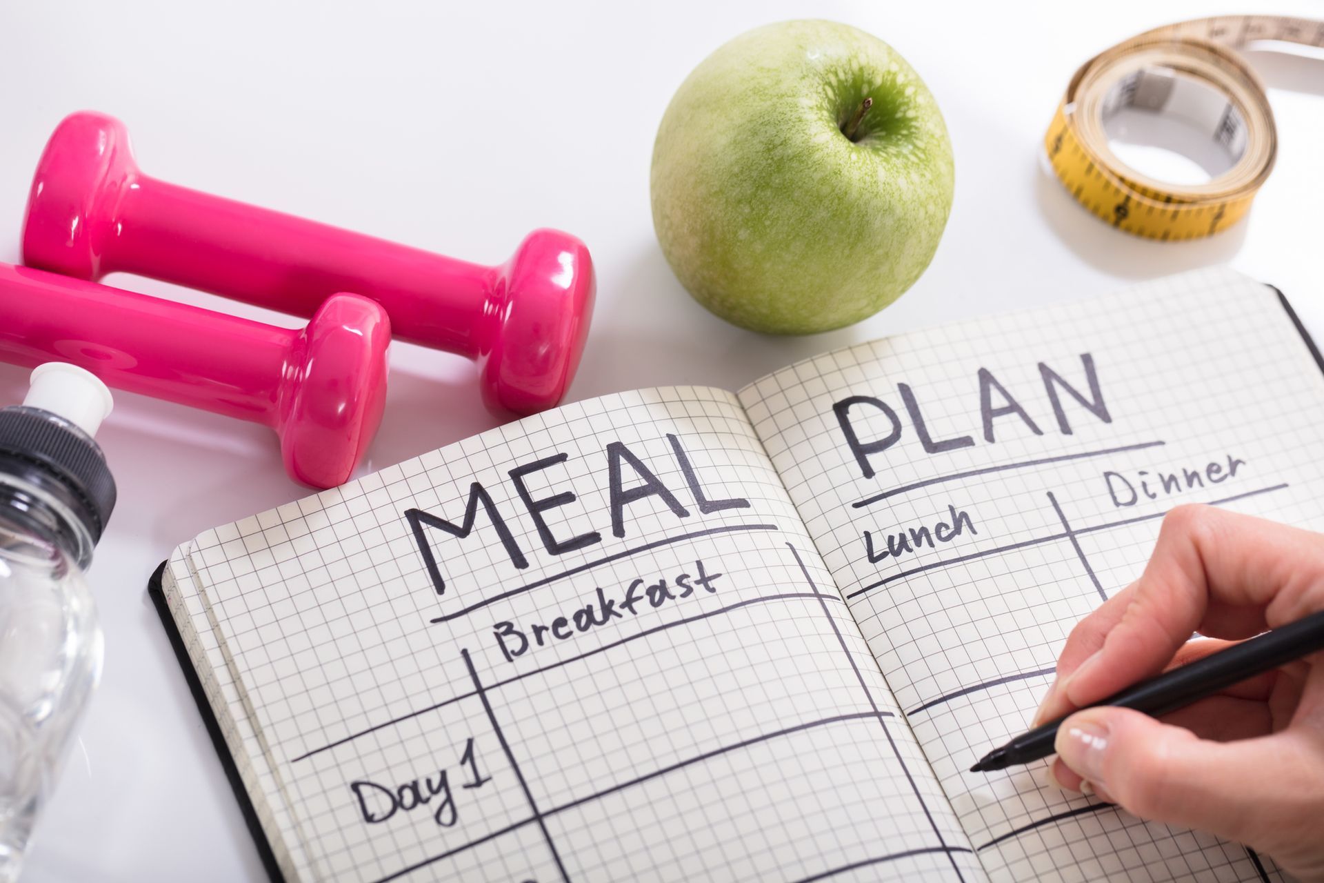 Person writing a meal plan in a notebook, alongside dumbbells, an apple, and measuring tape. Person writing a meal plan in a notebook, alongside dumbbells, an apple, and measuring tape.