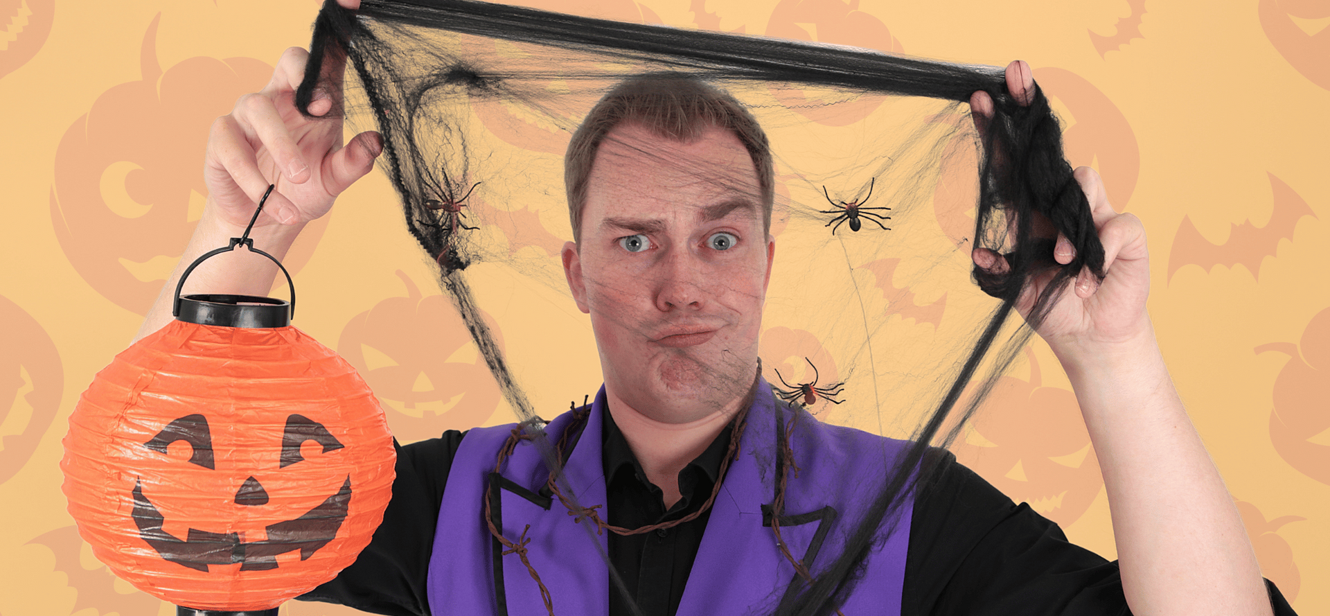 A man in a purple vest is holding a halloween lantern and a spider web.