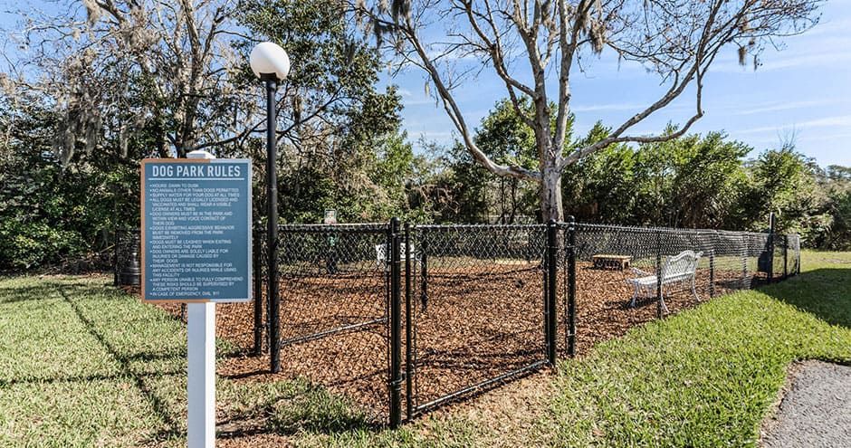 Fenced dog park with sign; green grass, brown mulch, trees, black metal fence, and a lamppost.