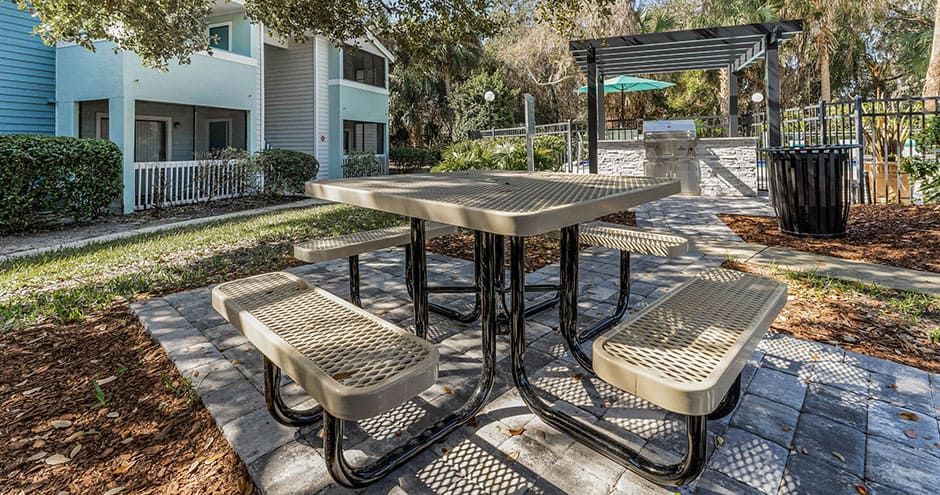 Picnic table with benches on a paved patio, near a grill, under a pergola, with a light blue apartment building.