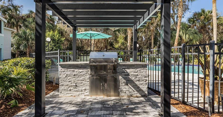 Outdoor kitchen with a grill under a pergola near a pool and greenery.