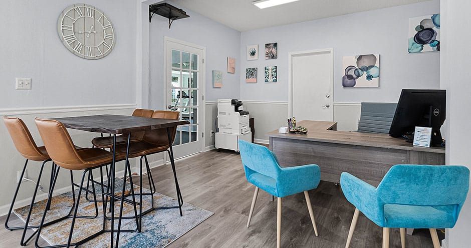 An office space with a table, chairs, desk, and decorations. Blue chairs at a desk, brown chairs around a table.