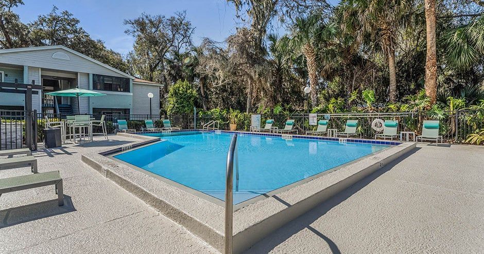 Swimming pool surrounded by lounge chairs, in front of a building and lush trees.