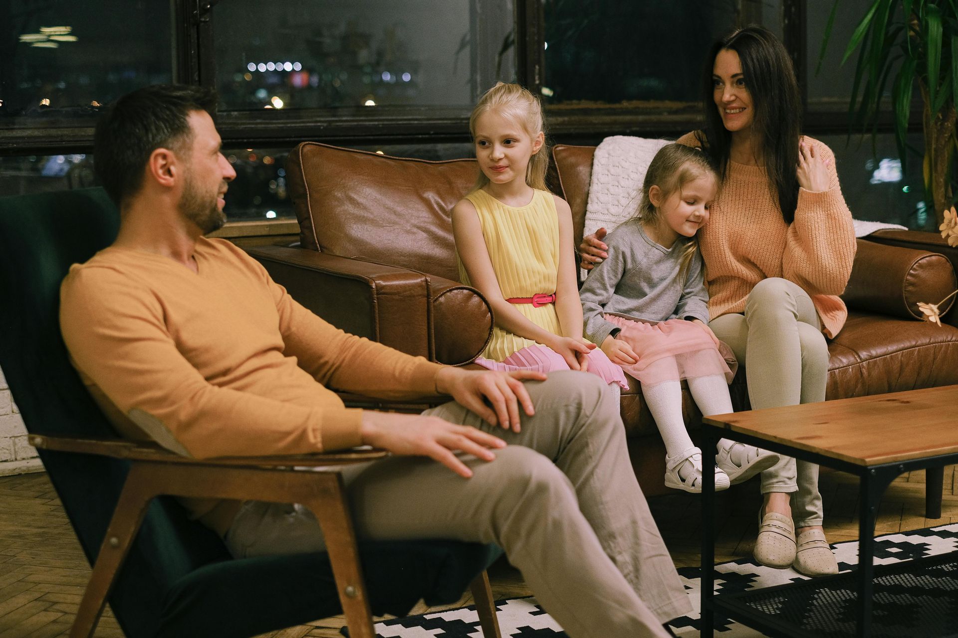 Family of four in living room, smiling; dad in chair, mom on sofa with two daughters.