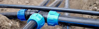 Black irrigation pipes connected by blue plastic fittings resting in a dirt trench.