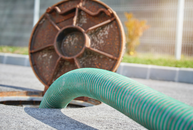 A green suction hose feeds into an open manhole on an asphalt road with a raised rusty metal cover in the background.