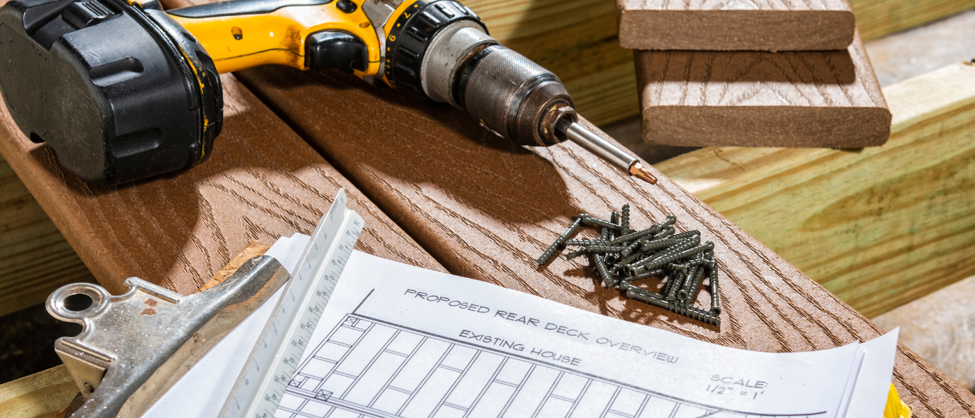 A photo of a drill, screws, and drawings for a deck plan. All of those items are sitting on composite deck boards, which are resting on pressure-treated wood.