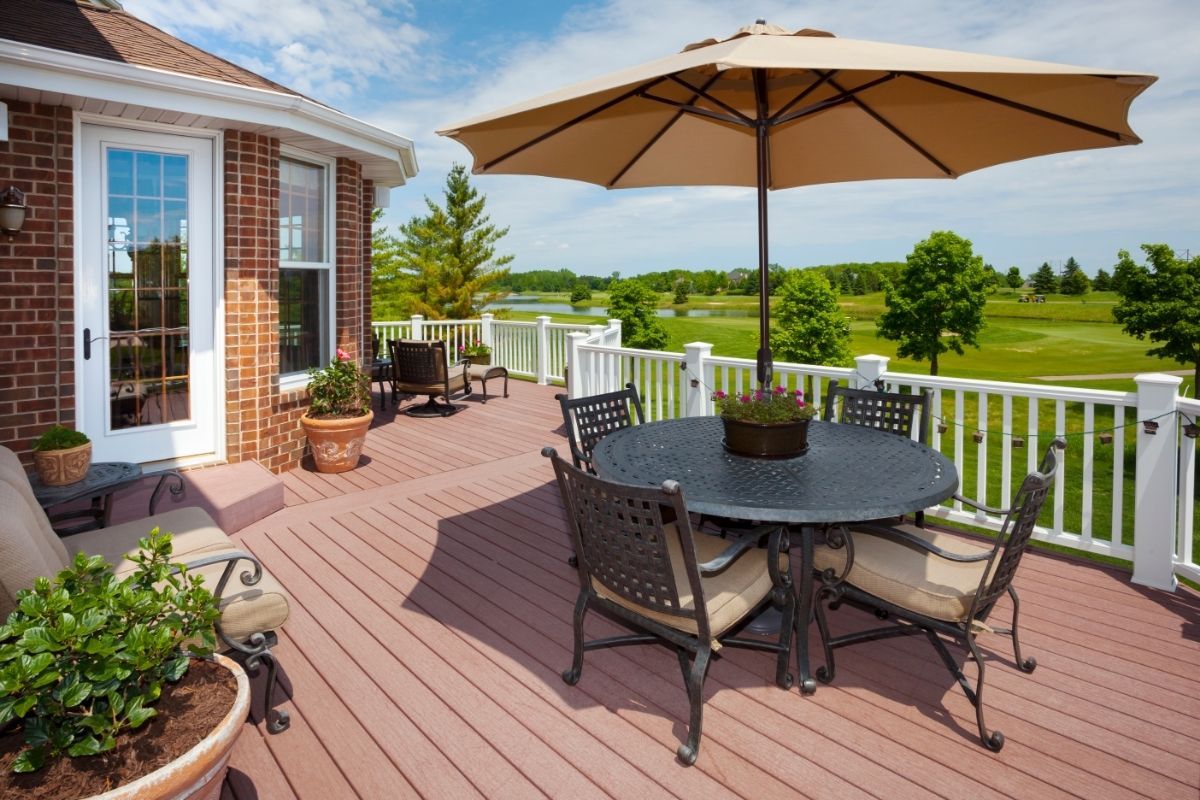 An expansive deck in Athens, GA, overlooks a lush golf course, offering a tranquil outdoor setting. The deck is equipped with a dark wrought iron table and chairs set, complemented by a large beige umbrella providing shade. Terra cotta planters with blooming flowers and verdant shrubs add a touch of color and life to the space. The deck’s reddish-brown planks contrast beautifully with the classic white railing and the home's brick exterior, creating an inviting space for relaxation or socializing.