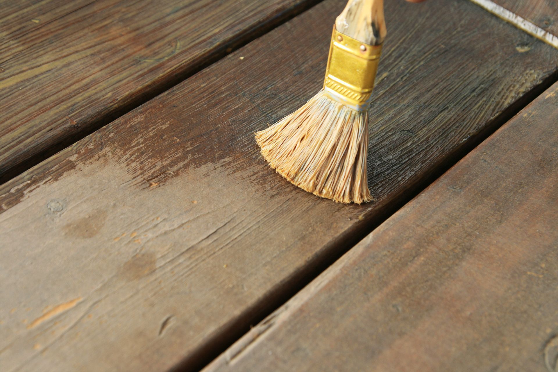 A used paintbrush with worn bristles rests on a partially stained wooden deck in Athens, Georgia, illustrating a work in progress. The old deck boards show signs of wear and previous paint layers, with a clear distinction between the unstained and freshly stained areas, demonstrating the renewing effect of the stain application.