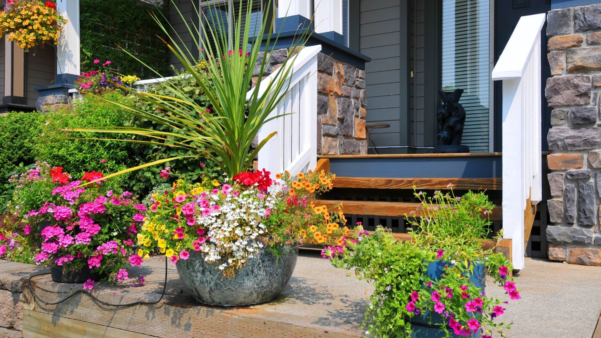 This vivid image captures a lush, vibrant garden at the front of a home in Athens, GA, showcasing a variety of colorful flowers in full bloom. The garden features a mix of pink, yellow, red, and white flowers, potted in large decorative containers and surrounded by lush greenery. A wooden deck with a small staircase leads up to the home's entrance, flanked by stone columns and a dark blue siding with white accents, creating a welcoming and picturesque scene.