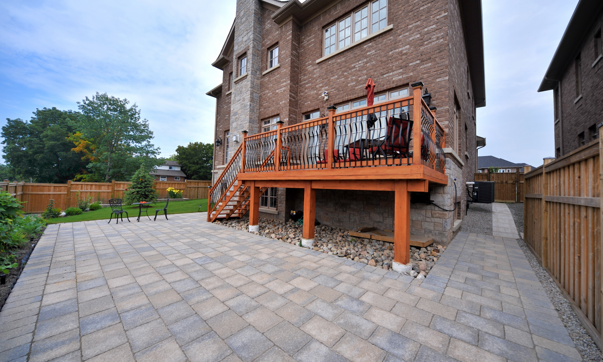 photo of a beautiful second-level deck on the back of a stunning house. The deck is a brown/orange color. There is a paver patio next to the deck.