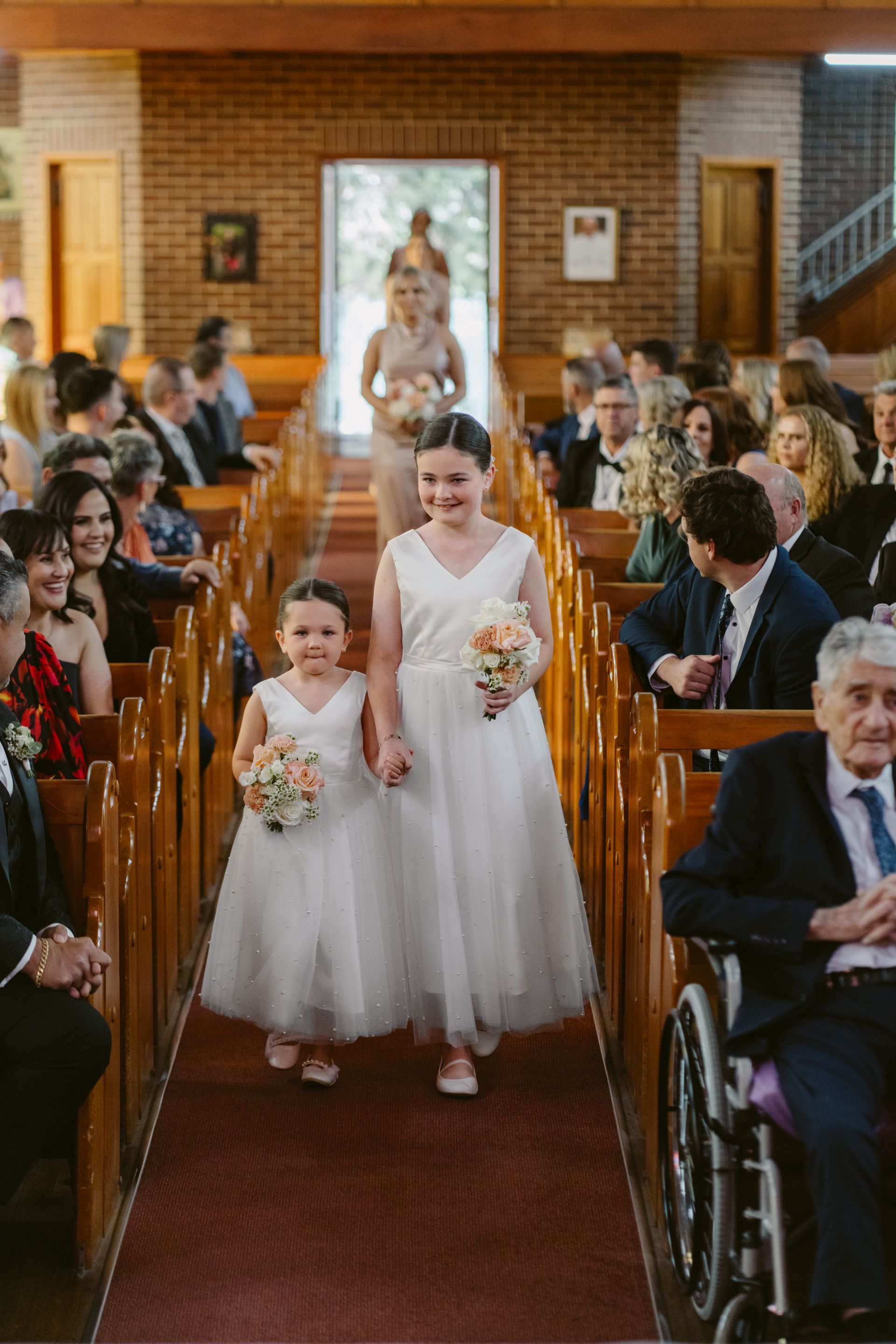 Two flower girls in white dresses walk down a church aisle towards the bride.