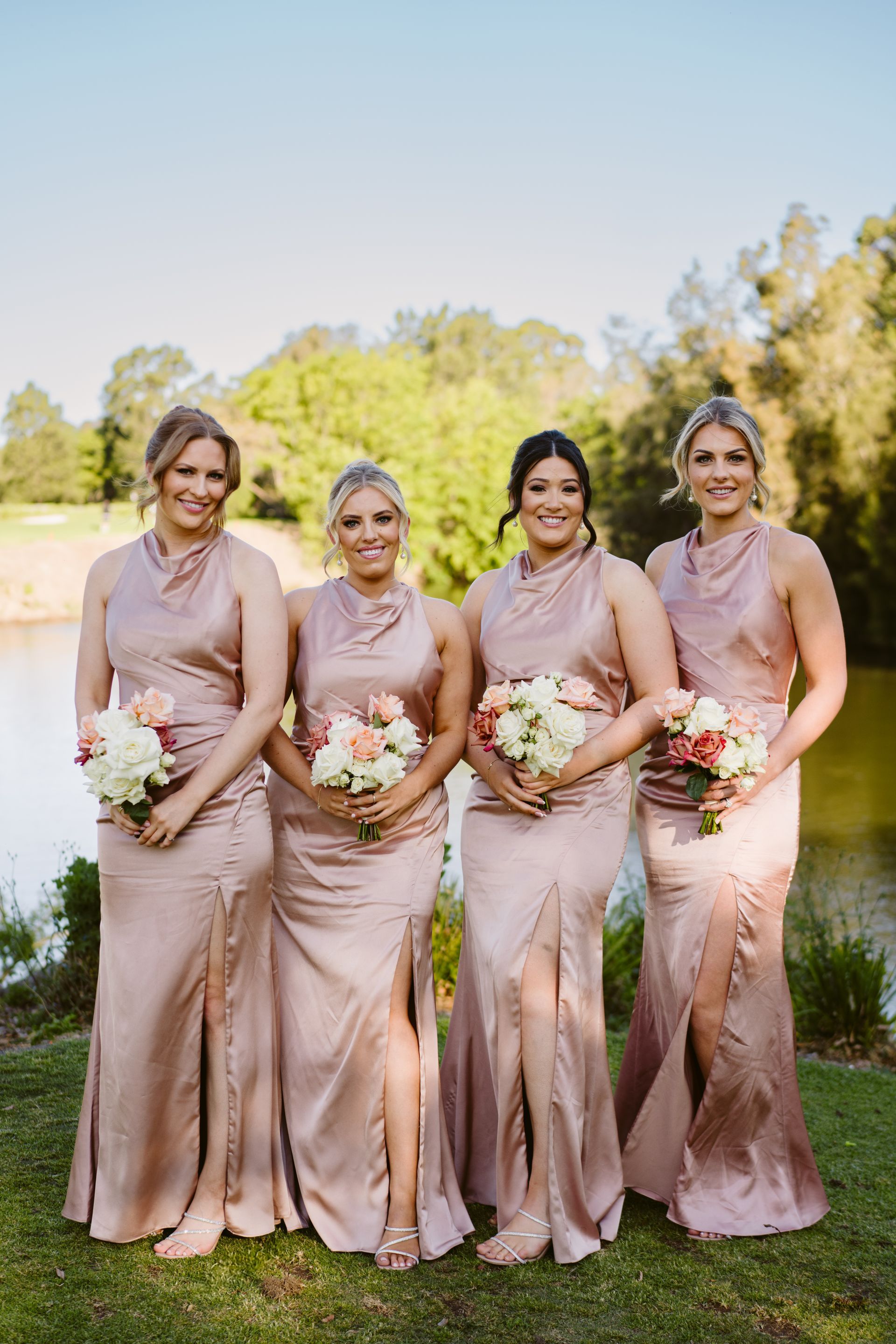 Four women in rose-colored dresses hold bouquets, standing outdoors near water, smiling.