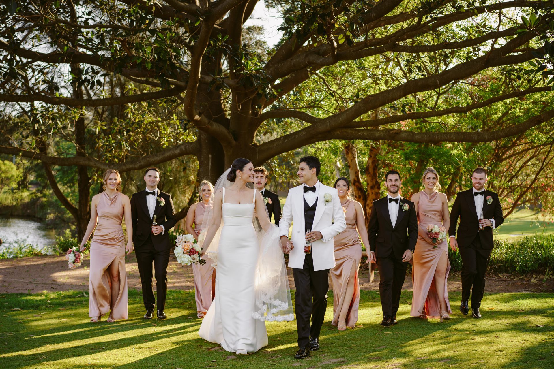 Wedding party walking on grass under a large tree, bride and groom in the center, bridesmaids in pink, groomsmen in black.