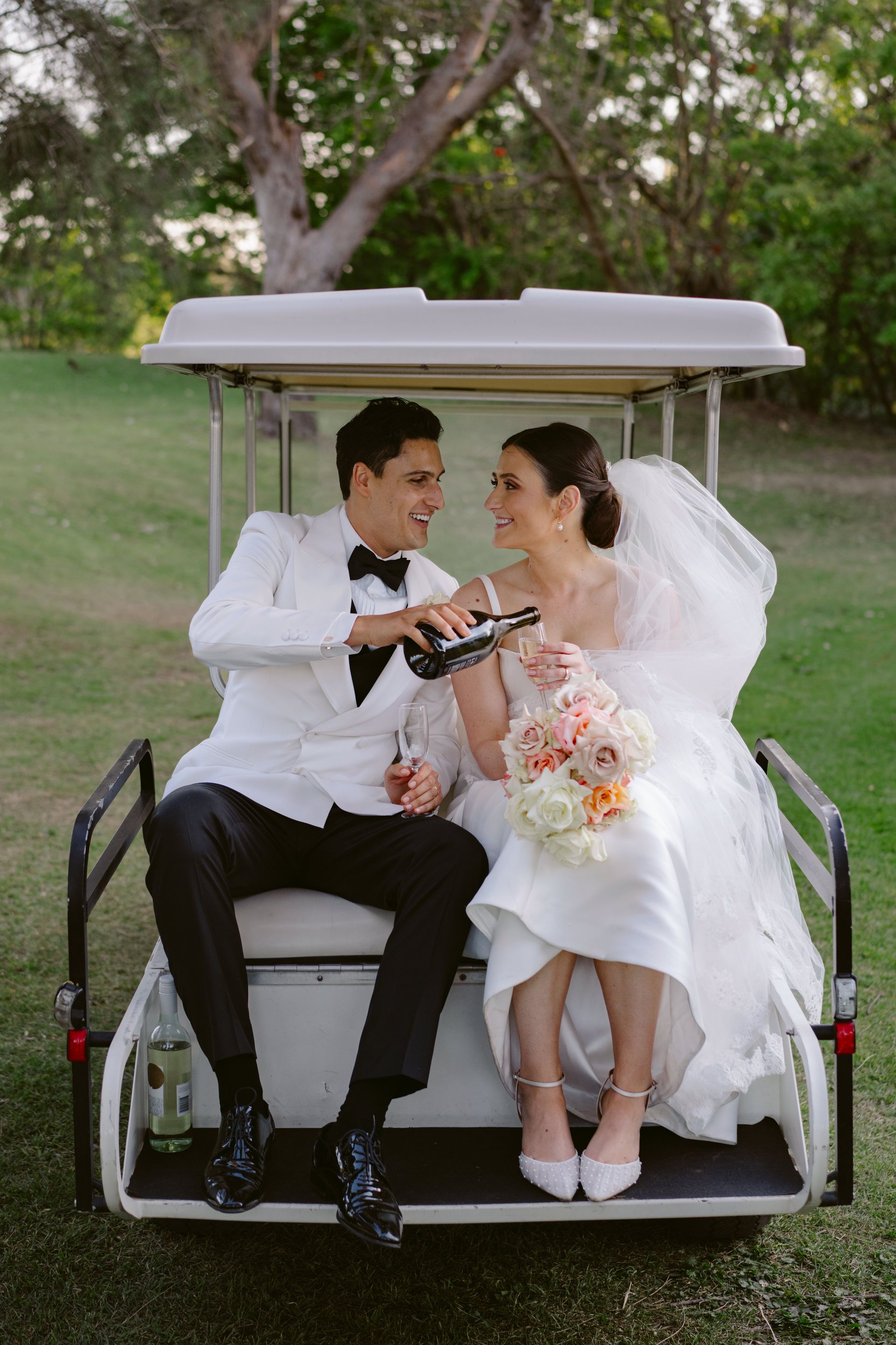 Bride and groom in golf cart, man pouring champagne. They are smiling, with a bouquet. Outdoors setting.