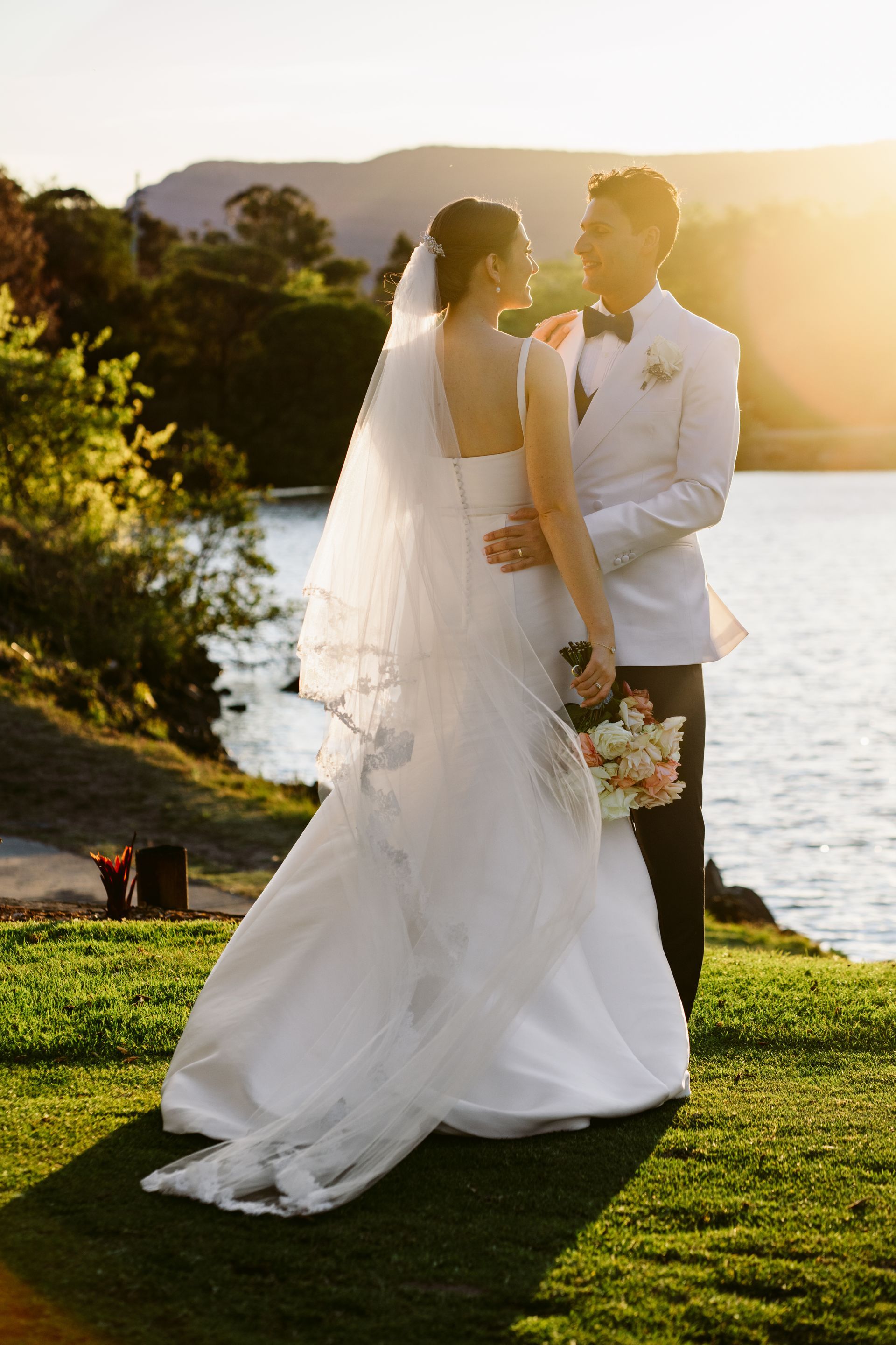 Bride and groom embracing, overlooking a lake at sunset. The bride wears a white gown, veil, and holds a bouquet.