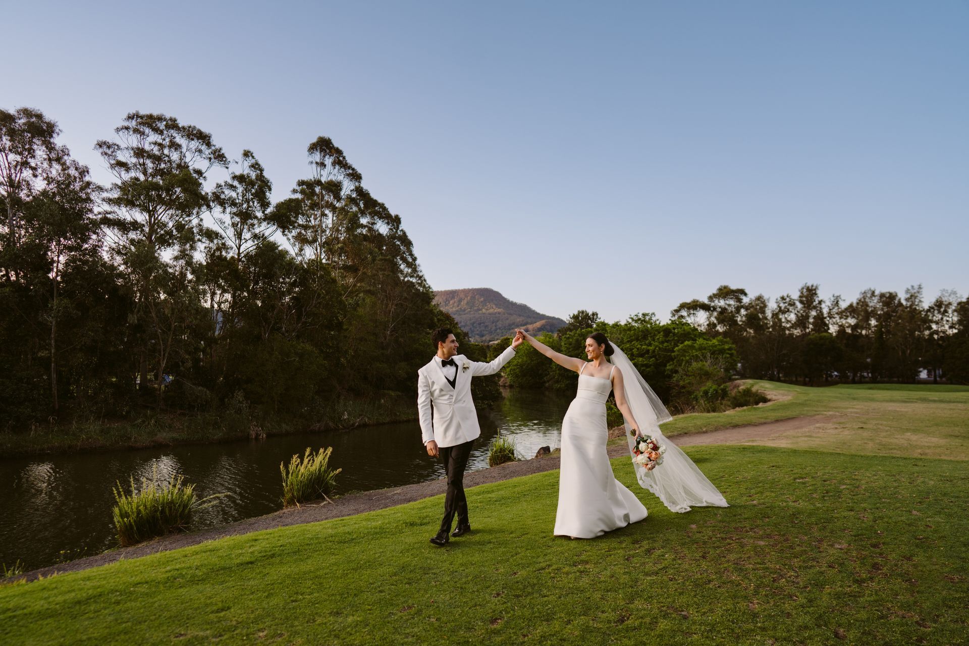 Newlyweds holding hands, dancing on a grassy bank near water, mountain backdrop, golden hour.