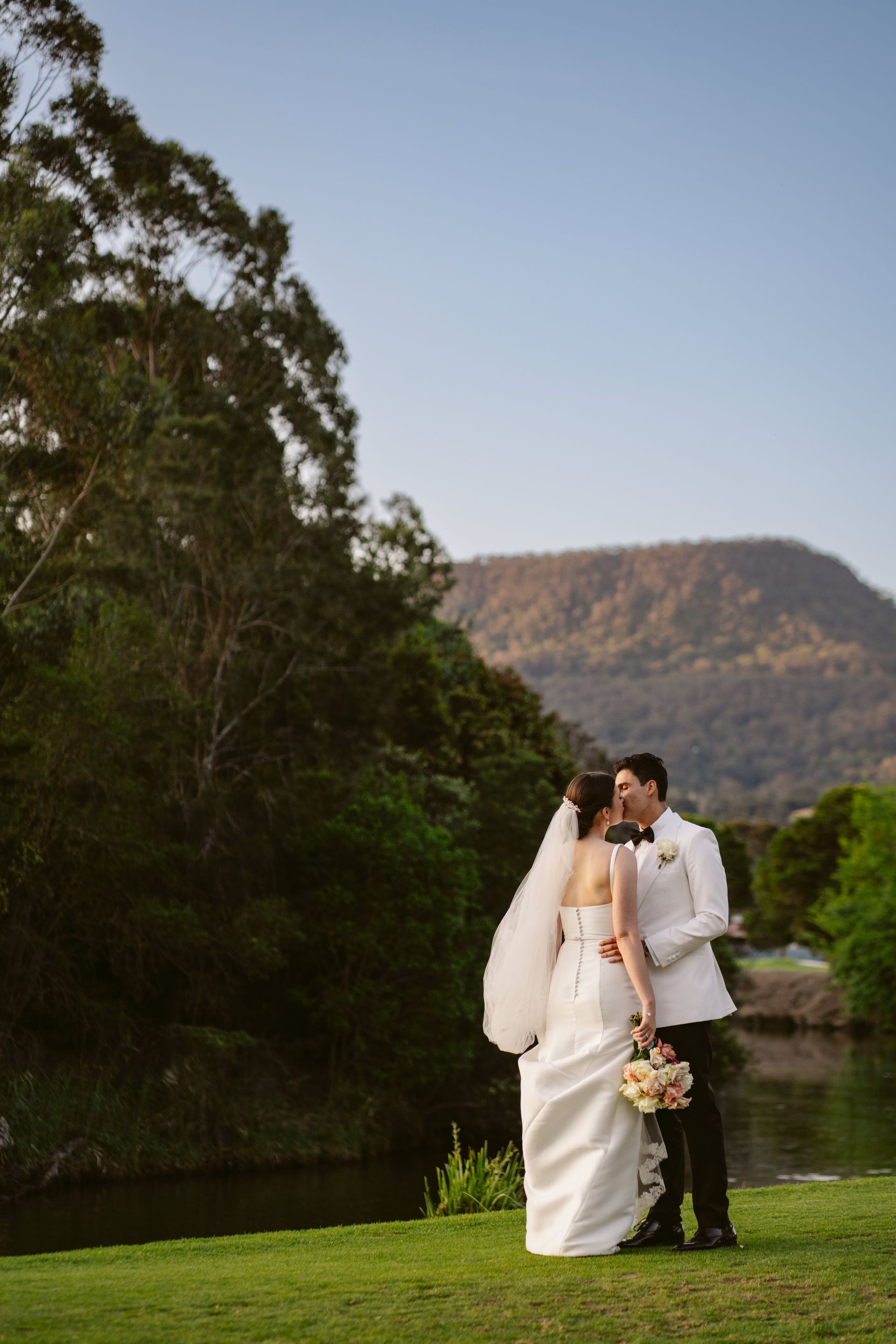 Wedding couple kissing, standing on green grass, overlooking a lake and mountains.