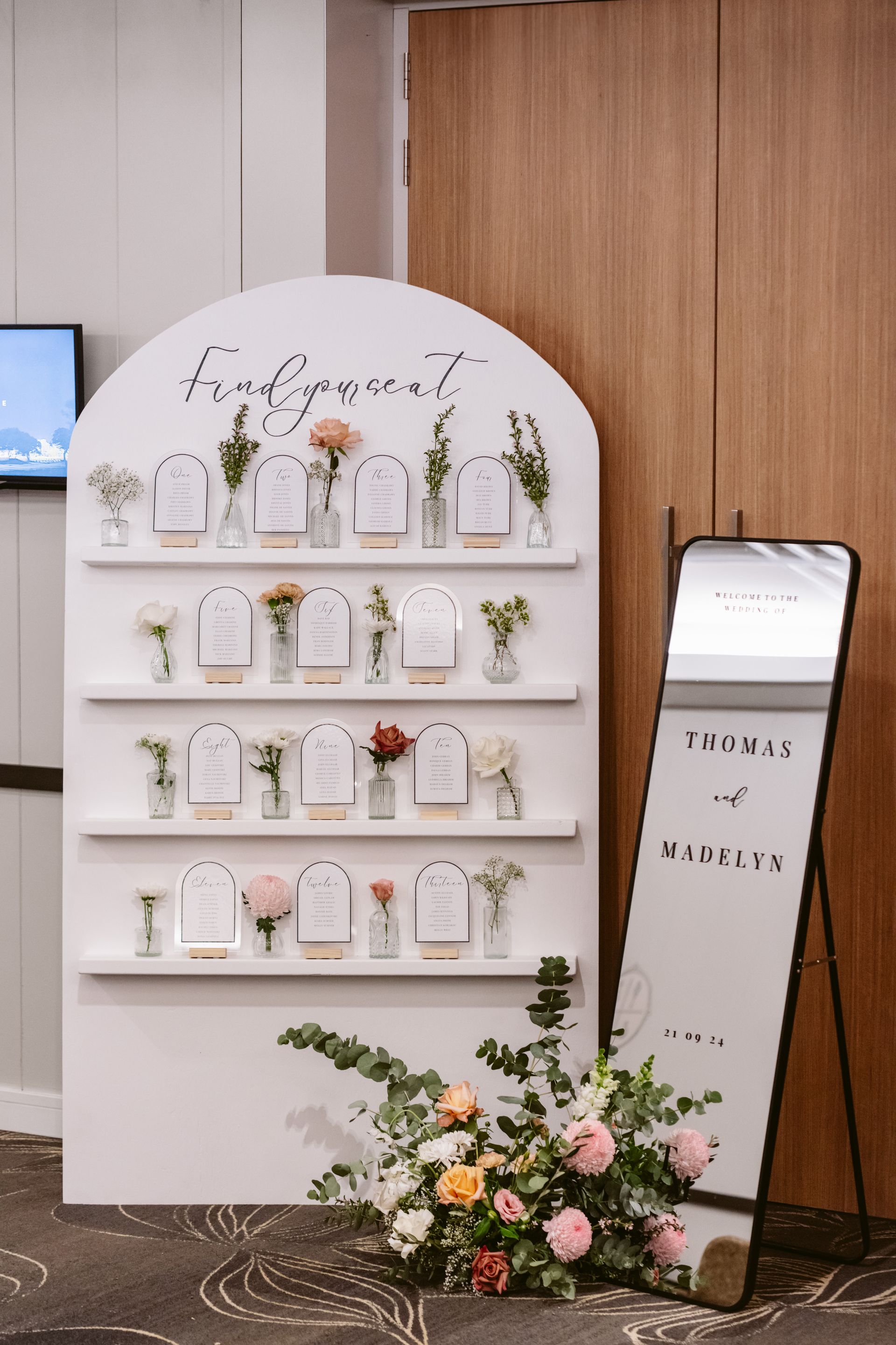 Wedding seating chart with floral arrangements, next to a mirror.