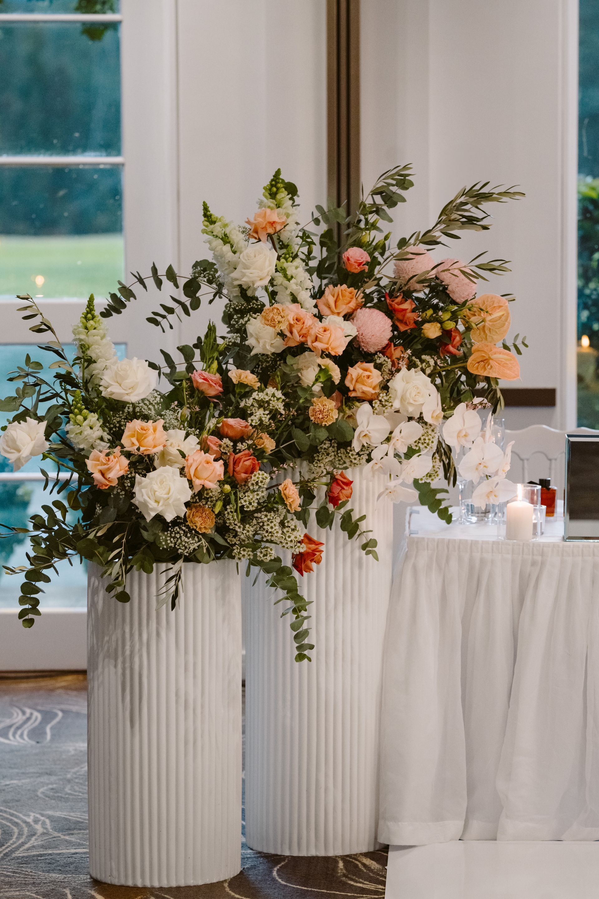 Two tall white floral arrangements with peach and white flowers on a table.